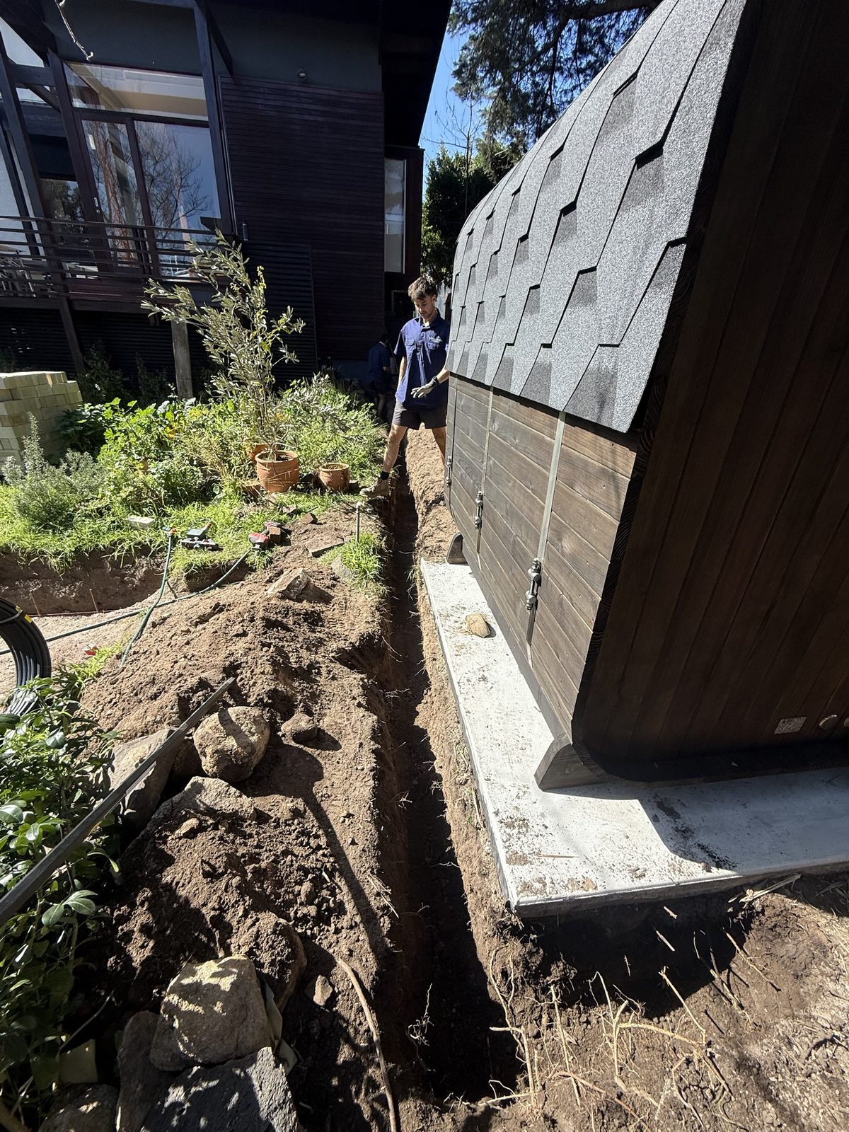 Licensed electrician inspecting underground three-phase cable trench alongside barrel sauna cabin Kew